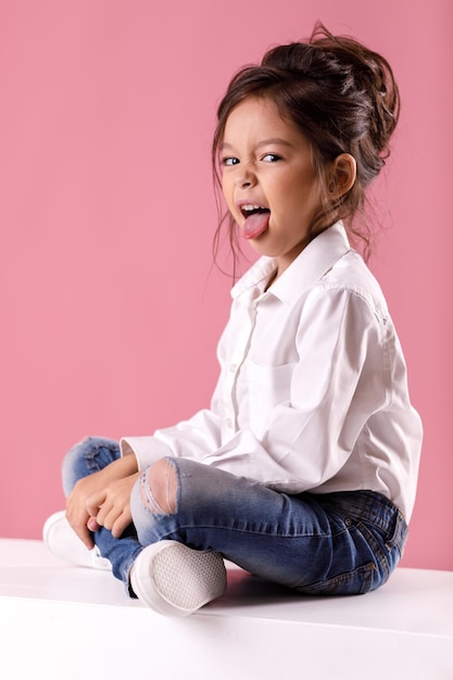 Cute little child girl in white shirt with hairstyle showing tongue on pink background. Human emotions and facial expression