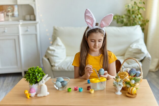 バニーの耳をかぶっているかわいい小さな子供。リビングルームのテーブルに座って座っている女の子。子供は卵を描いています。
