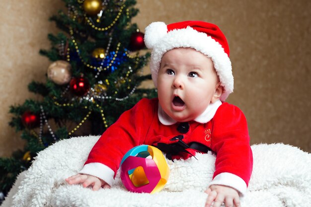 Cute little christmas baby wearing santa claus suit with a funny surprised look , christmas tree at the wall