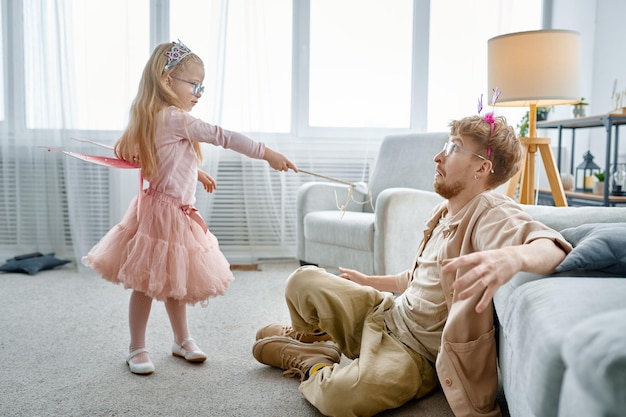 Cute little daughter princess with magic wand and father wearing costumes having fun at home