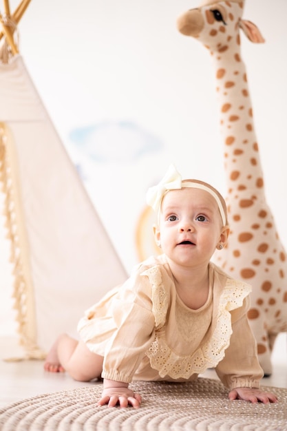 A cute little European girl with brown eyes in a beige bodysuit made of natural fabric plays in the children's room at home against the background of a wigwam and plush toys