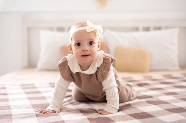 A cute little European girl with brown eyes in a beige knitted suit crawls on the bed at home smiles home textiles