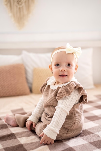 A cute little European girl with brown eyes in a beige knitted suit is sitting on the bed at home smiling home textiles