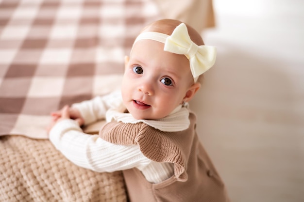 A cute little European girl with brown eyes in a beige knitted suit is standing by the bed at home smiling a closeup portrait of a child