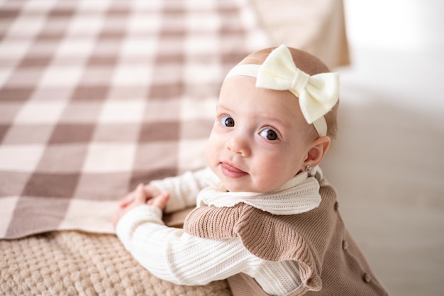 A cute little European girl with brown eyes in a beige knitted suit is standing by the bed at home smiling a closeup portrait of a child