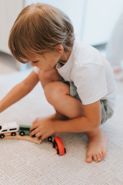 Cute little five year old kid boy in a white t-shirt playing with a wooden railway and toy trains on the floor on the carpet in the room. children plays at home