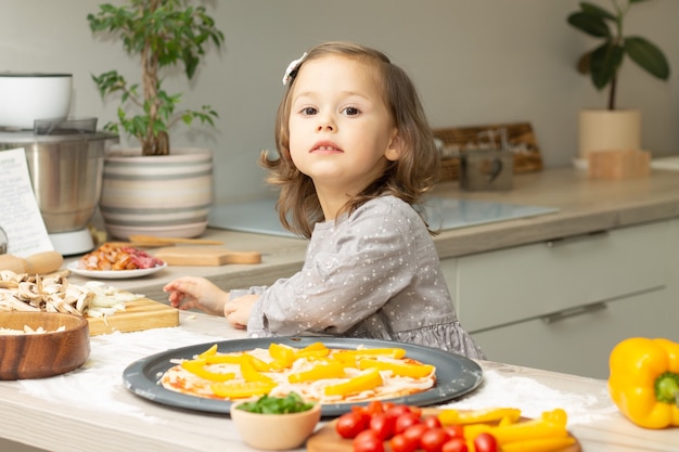 写真 キッチンでピザを調理する灰色のドレスを着たかわいい女の子2-4。子供はピザベースに材料を配置します