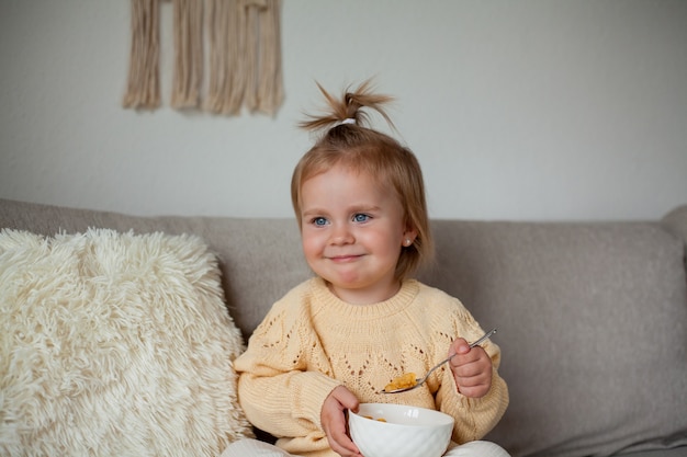 A cute little girl 2 years old in a cozy knitted sweater is having breakfast on the couch