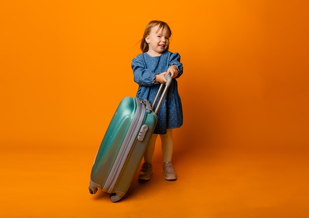 Cute little girl 4 years old in a blue denim dress with a green suitcase on a yellow background.