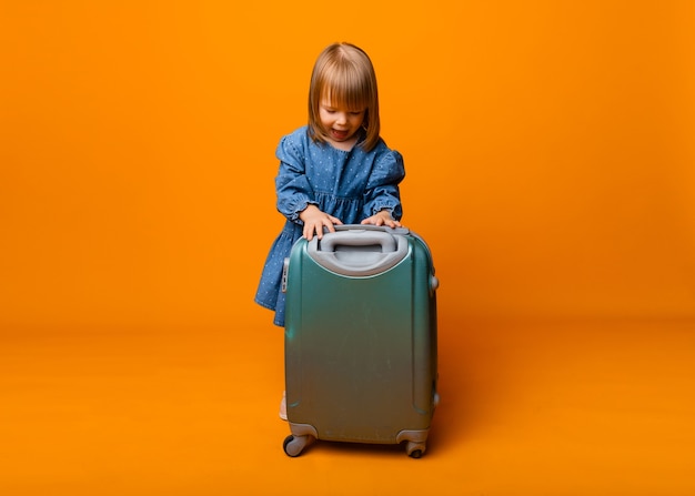 Cute little girl 4 years old in a blue denim dress with a green suitcase on a yellow background.