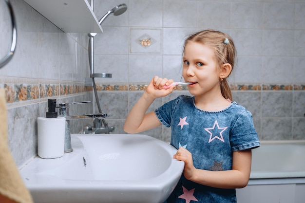 A cute little girl of 5 years old looks at the camera with a slight smile and brushes her teeth in the bathroom