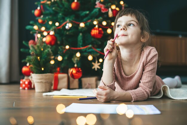 Cute little girl 56 years old writing her wishes to Santa by the festive tree her eyes filled with dreams