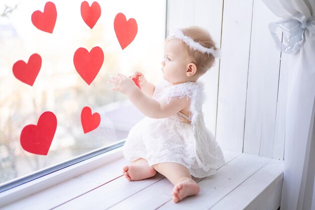 A cute little girl in an angel costume with wings is sitting on the windowsill near the window against a background of red hearts valentines day concept valentines day