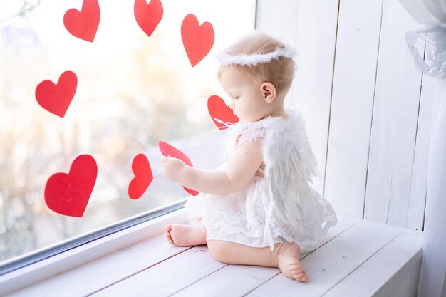 A cute little girl in an angel costume with wings is sitting on the windowsill near the window against a background of red hearts valentines day concept valentines day