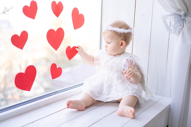 A cute little girl in an angel costume with wings is sitting on the windowsill near the window against a background of red hearts valentines day concept valentines day
