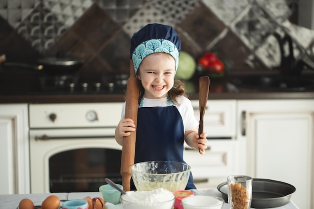 Una ragazzina carina in grembiule e cappello da chef sta appiattendo l'impasto usando un mattarello