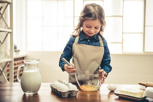 Cute little girl in apron is smiling while whisking eggs for baking in the kitchen