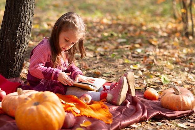 Cute little girl in autumn park with books and orange color leaves and yellow pumpkin