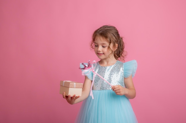 A cute little girl in a beautiful dress holds a fairy's magic wand and a gift on a pink background