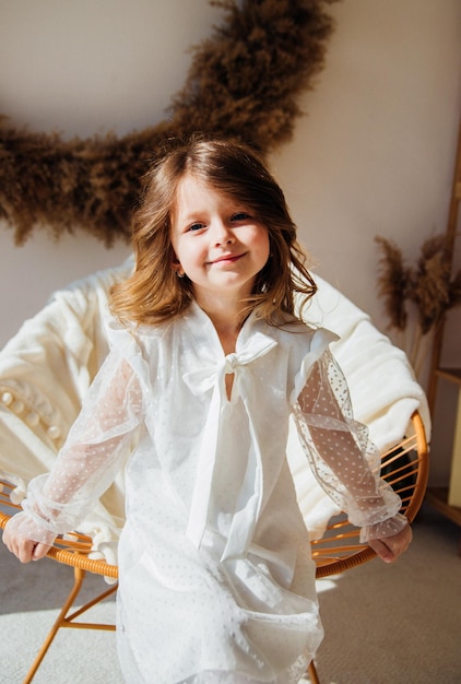 A cute little girl in a beautiful dress is sitting in a chair in a bright sunny room Sunlight falls from the window