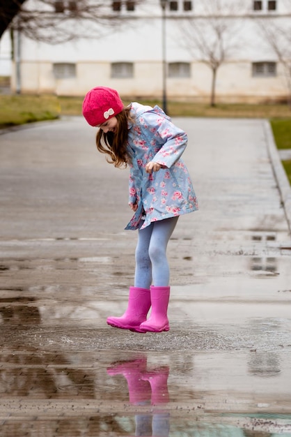 파란색 망토, 분홍색 부츠, 분홍색 모자를 쓴 귀여운 소녀가 웅덩이를 뛰어다니며 즐겁게 놀고 있습니다.
