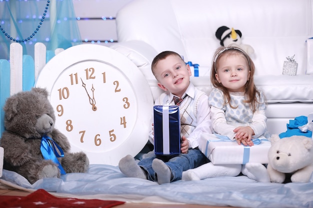 Cute little girl and boy with gifts boxes and big white clock sitting near the white sofa in Christmas and New Year