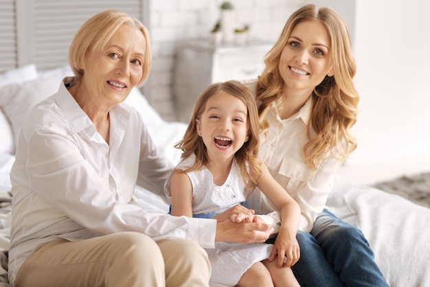 Cute little girl bursting into laughter while sitting in between her charming mother and grandmother holding hands together around her