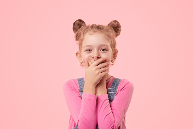 Cute little girl in casual clothes covering mouth with hands while keeping secret and looking at camera with smile against pink background