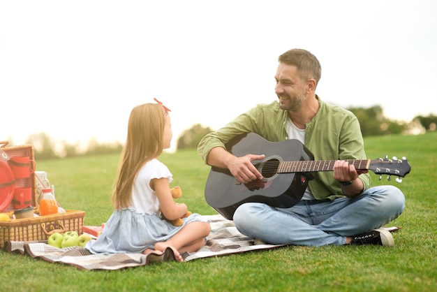 Cute little girl in casual clothes sitting on a green grass in park and looking at her loving dad