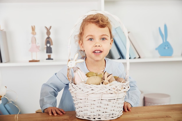 Cute little girl celebrating Easter holidays indoors near table with basket with eggs