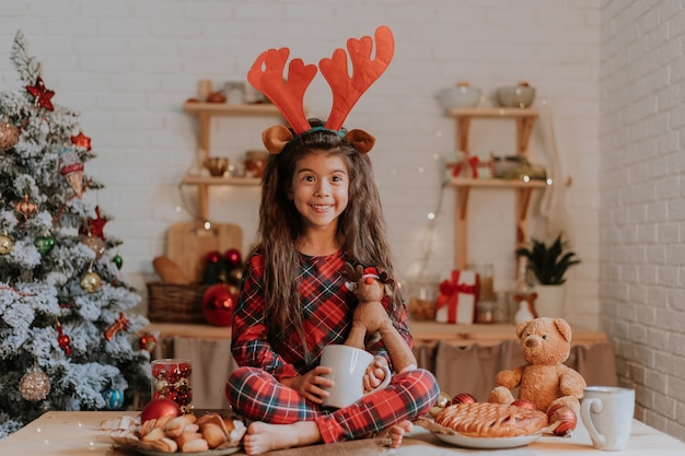 Cute little girl in checkered red pajamas with reindeer horns on her head is eating a Christmas cake