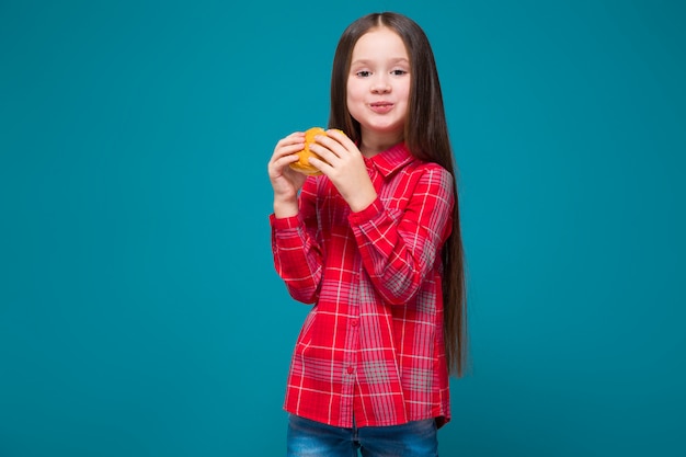 La bambina sveglia in camicia a quadretti con i capelli della brunetta tiene l'hamburger