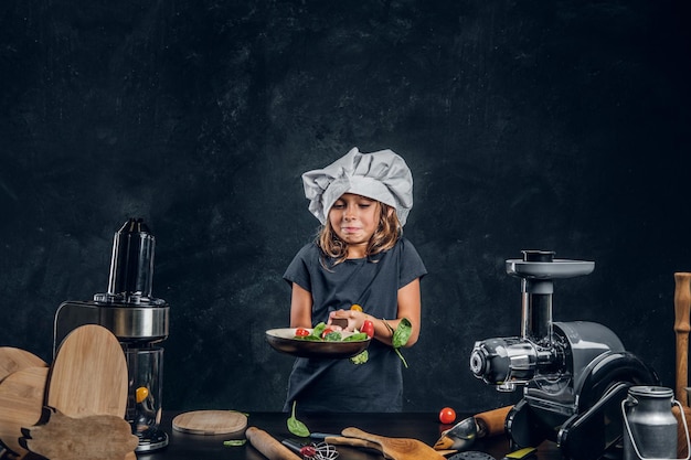 Cute little girl in chef's hat is preparing vegetables for cooking on the dark background.