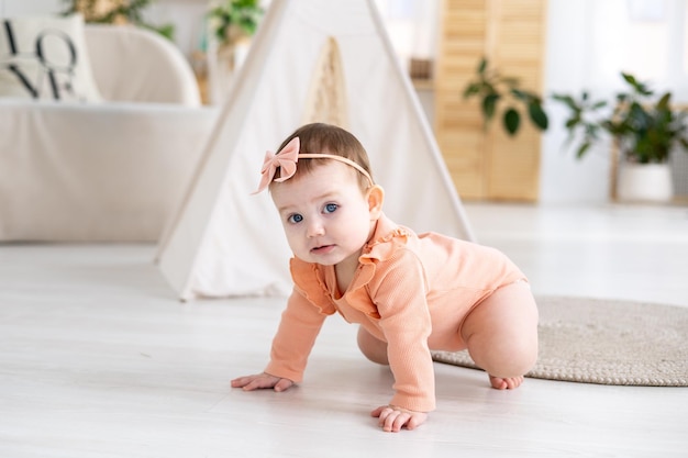 Photo a cute little girl child in a pink bodysuit crawls on a rug in the living room of the house against the background of a wigwam looks at the camera smiles