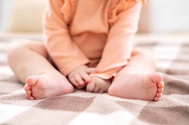 A cute little girl child in a pink bodysuit is sitting on a bed on white bedding with a beige plaid at home in the bedroom naked baby legs closeup baby toes