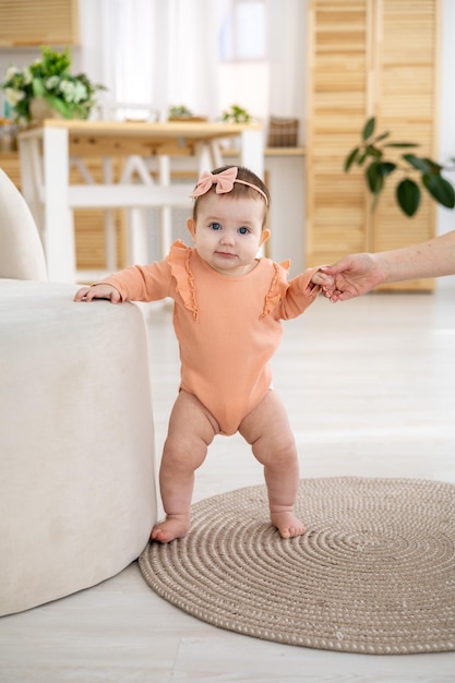 A cute little girl child in a pink bodysuit is standing in the living room of the house holding on to the support the first steps of the child the baby is learning to walk