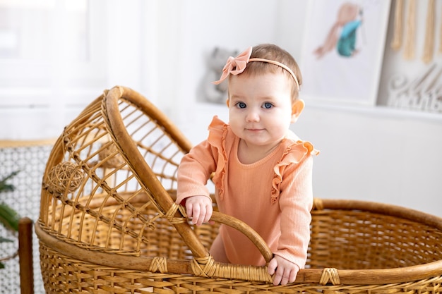 A cute little girl child in a pink bodysuit is standing in a wicker cradle at home looking at the camera smiling children's healthy sleep