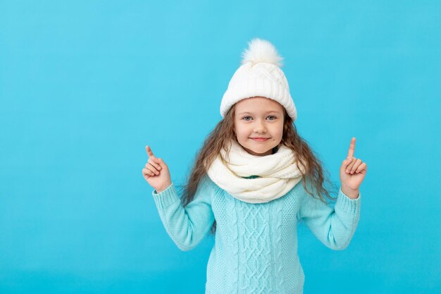 Cute little girl child in winter clothes hat and sweater points fingers up at something on a blue isolated background and smiles and laughs, a place or space for text