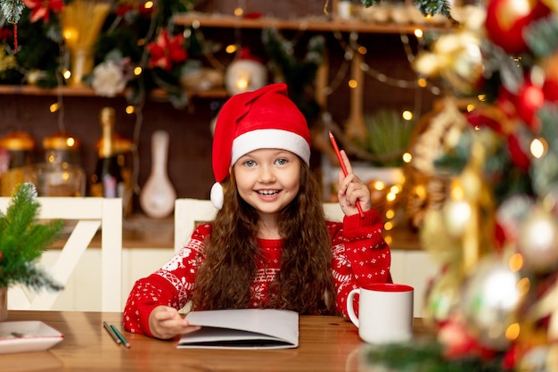 A cute little girl child in winter clothes a Santa Claus hat and a red sweater writes a letter to Santa Claus in the dark kitchen of the house and smiles and waits for the new year and Christmas