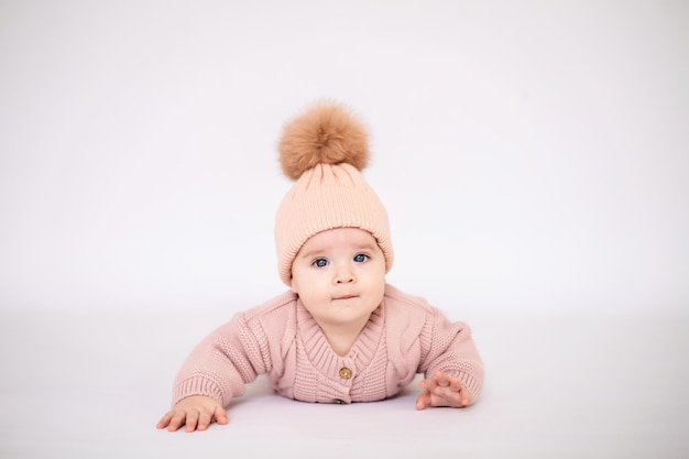 A cute little girl child with blue eyes in a pink knitted jumpsuit and a hat lies on her stomach looking at the camera on a white background isolated