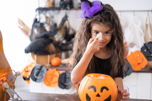 Photo cute little girl in costume of witch holding pumpkin jack with candies, having fun in kitchen, celebrating halloween.