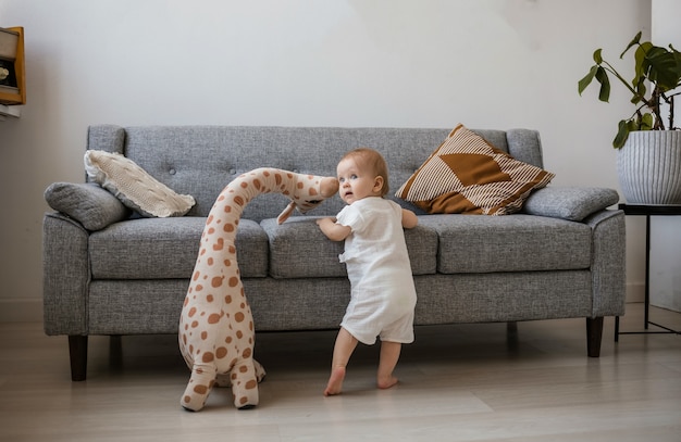 A cute little girl in a crimson jumpsuit is standing next to a gray sofa with a stuffed giraffe toy in the room