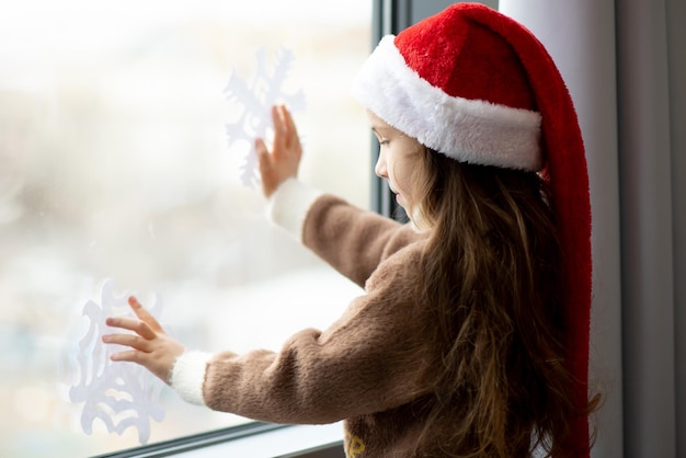 귀여운 소녀가 눈송이로 창문을 장식합니다. 새해 크리스마스 분위기
