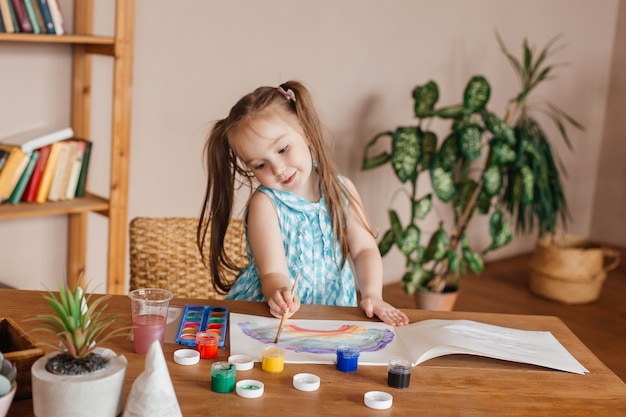 Cute little girl draws with a brush and paints at the table in the living room