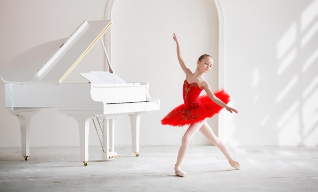 プロのバレリーナになることを夢見ているかわいい女の子ピアノの隣の白い部屋で、真っ赤なチュチュの女の子がトウシューズで踊っています専門学校の学生
