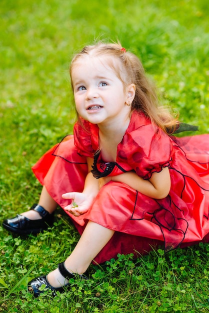 Милая маленькая девочка, одетая в длинное красное платье, играет и веселится на зеленой лужайке