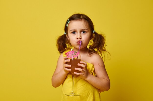 Cute little girl drinking a tropical fruit cocktail from a straw, isolated on yellow with copy space.