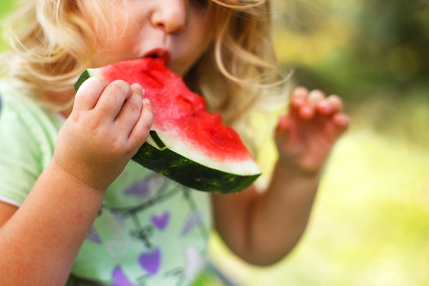 Cute little girl eating a piece of watermelon close in the garden in summertime