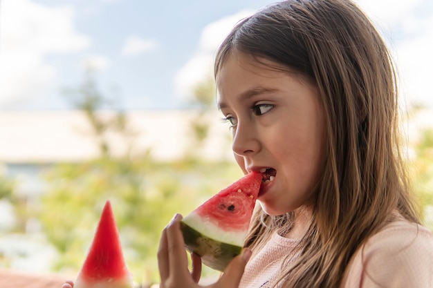 夏にスイカを食べるかわいい女の子。子供は熟した赤いジューシーなスイカを噛みます。