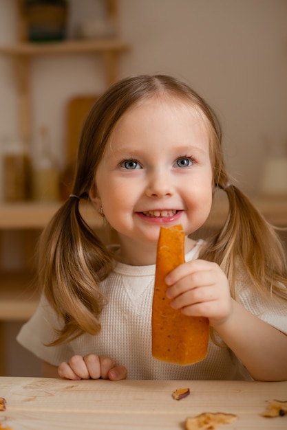 Cute little girl eats natural pastille at home in a wooden kitchen.